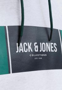 Hættebluse med hvid krop, der har en rektangulær sort og teal grafik samt en grøn flettet snor med sorte spidser.