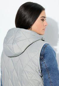 Hellgraue gesteppte Weste mit Kapuze; verfügt über Denim-Ärmel und eine glatte Textur. Sichtbare Nähte an der Weste und Denim-Akzente.