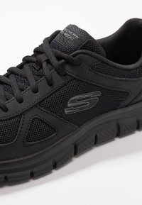 Black athletic shoe featuring a mesh upper, padded collar, and textured rubber sole with a visible "S" logo on the side. Lace-up design.