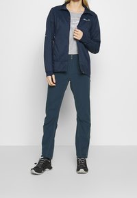 Navy lightweight jacket with zip front, paired with teal pants and black athletic shoes. Grey T-shirt features subtle graphic design.