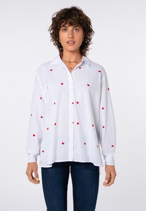 Frau mit lockigem Haar, die ein weißes, langärmeliges Hemd mit kleinen roten Herzmustern und dunkelblaue Jeans trägt.