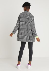 Blazer oversize a quadri bianco e nero, maniche lunghe e polsini in un verde chiaro a contrasto, indossato sopra jeans skinny scuri e sneaker bianche.