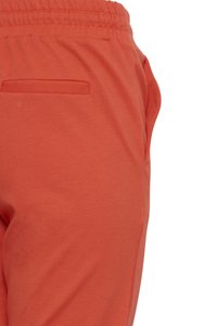Detail oranžových nohavíc s elastickým pásom, zadným límcom vrecka a viditeľným bočným švom na bielom pozadí.