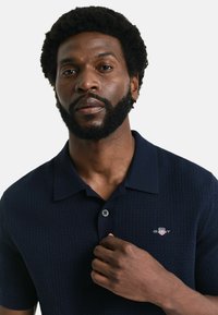 Marinblå pikétröja med en texturerad design, tvåknappslåstning och en liten broderad logotyp på bröstet. Kortsleeves.