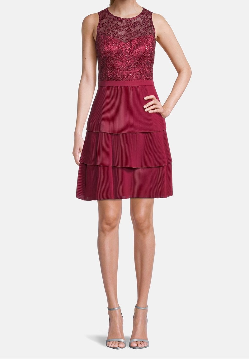 Robe bordeaux sans manches avec corsage en dentelle et jupe plissée à étages, portée avec des sandales à talons hauts argentées à bride cheville.