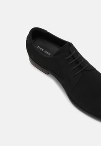Chaussure habillée en daim noir avec un bout arrondi, cinq œillets pour les lacets et un subtil accent rouge sur la semelle. L'intérieur présente une semelle intérieure de marque.