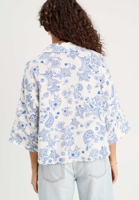Blouse blanche avec un motif paisley bleu, dotée d'un col et de larges manches. Le tissu semble léger avec une finition texturée.