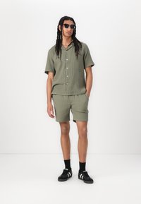 Camisa de manga corta de color verde oliva con rayas y botones, acompañada de pantalones cortos a juego. Gafas de sol negras y zapatillas negras con detalles blancos completan el conjunto.