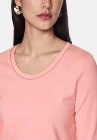Helle pinkfarbene Langarmbluse mit einem Rundhalsausschnitt und dezenten Nähten. Dazu große goldene kreisförmige Ohrringe.