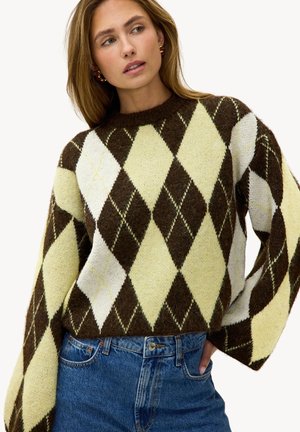 Joven mujer que lleva un suéter de rombos en marrón, crema y amarillo con jeans azules, posando con la mano en el bolsillo y mirando hacia un lado.