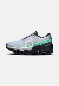 Chaussure de course gris clair avec une tige texturée, des accents noirs et des inscriptions vertes. Elle présente une semelle épaisse unique avec des découpes pour l'amorti.