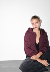 Mladá žena s hladko zloženými vlasmi, oblečená v marónovej mikine a čiernom oblečení, sedí so skríženými nohami na šedej podlahe pri bielej stene.