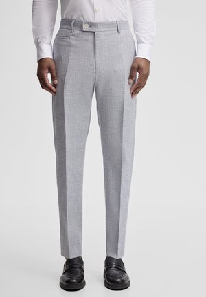 Man draagt lichtgrijze op maat gemaakte broek, wit overhemd en zwarte leren loafers, staand tegen een effen achtergrond.