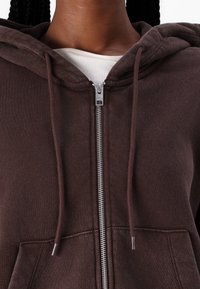 Felpa con zip di colore marrone, realizzata in tessuto morbido, dotata di cappuccio, cordini, tasche anteriori e dettagli con zip in tono argento.