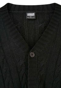 Gilet en tricot noir avec un décolleté en V, présentant des motifs en torsades et des côtes, fermeture par un bouton marron unique, et une étiquette indiquant "Urban Classics, M."