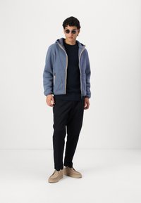 Blue fleece zip-up jacket with contrasting light inner lining, paired with a navy sweater and black trousers. Light brown shoes complete the look.