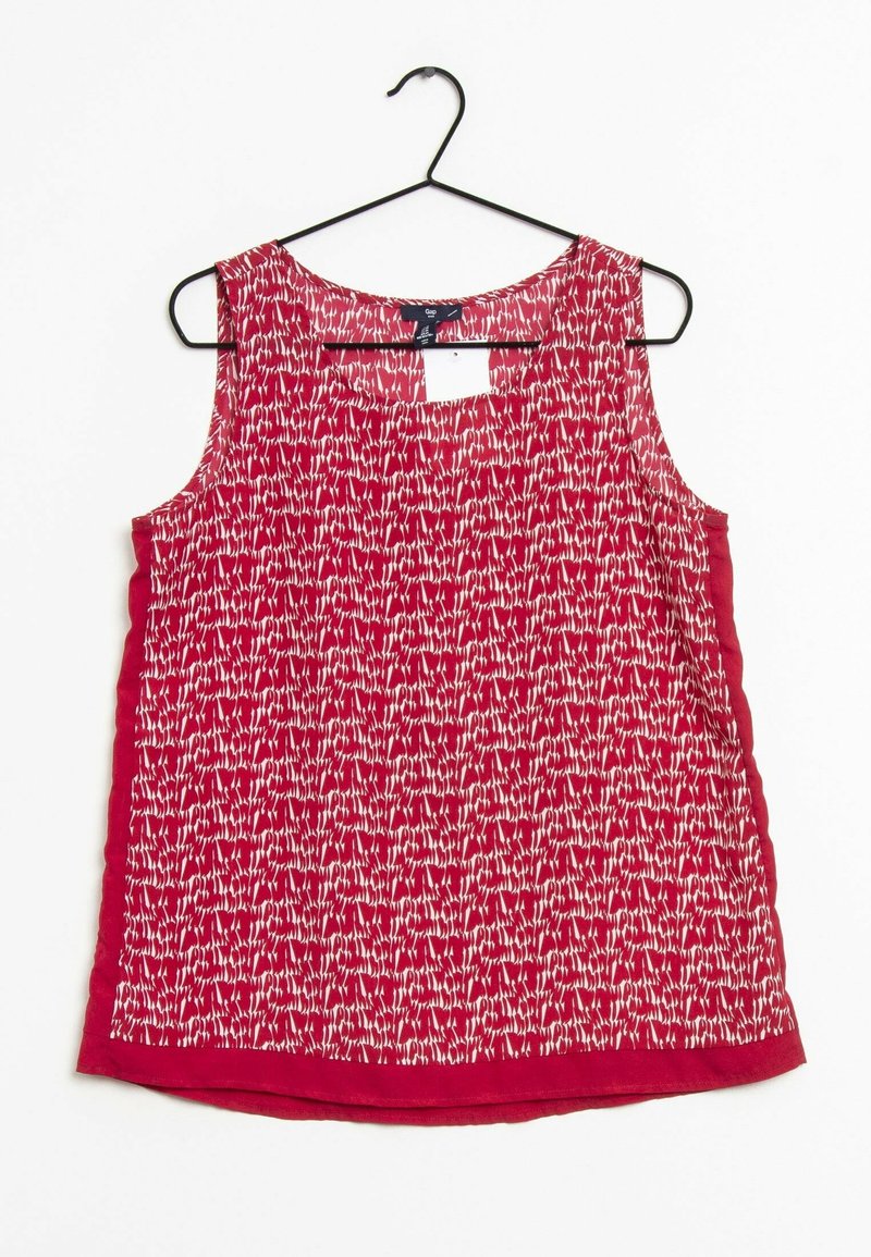 Blouse rouge sans manches avec motif abstrait blanc suspendue sur un cintre noir sur fond blanc.