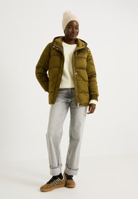 Doudoune vert olive avec une capuche, fermeture éclair et un design légèrement gonflé. Portée par-dessus un pull crème et un jean gris. Baskets marron.