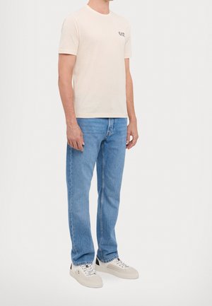 Uomo che indossa una t-shirt crema EA7, jeans dritti azzurri chiari e sneakers bianche, in piedi davanti a uno sfondo semplice.
