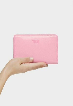 Cartera de piel rosa con textura suave, con un logo "TOUS" en relieve en la parte frontal, forma rectangular y cierre de cremallera.
