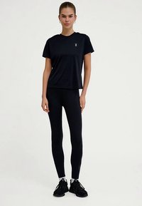 Svart atletisk t-shirt med korta ärmar och logotyp, kombinerad med svarta leggings. Båda plaggen är åtsittande och gjorda av stretchigt tyg.