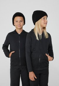 Des sweat-shirts à capuche noirs zippés et un pantalon assorti en tissu doux, avec des poches avant. Tous deux portent des bonnets en tricot noirs.