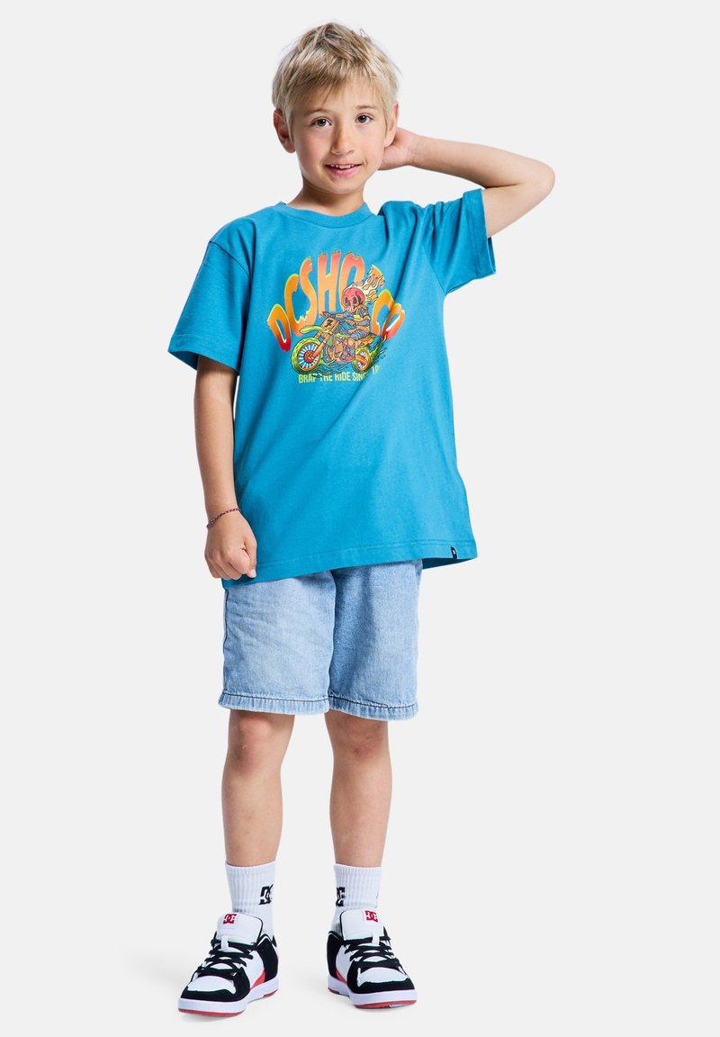 T-shirt en coton bleu vif avec un graphisme coloré, associé à un short en denim clair et des baskets noir et blanc.