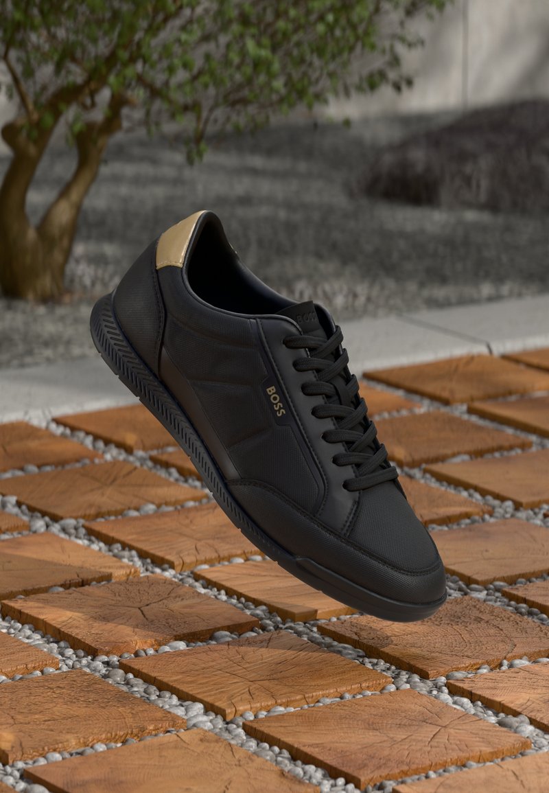 Black sneaker with a textured upper, gold accent at the heel, and laces. Rounded toe and flexible sole. Set against patterned tile background.