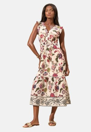 Frau trägt ein ärmelloses, V-Ausschnitt, knöchellanges Blumenkleid mit Rüschendetails, kombiniert mit goldenen flachen Sandalen.