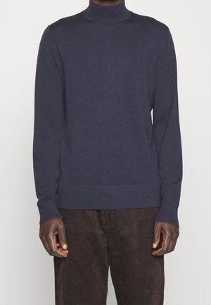 Homme portant un pull à col roulé bleu marine et un pantalon en velours côtelé marron foncé, debout devant un fond blanc.