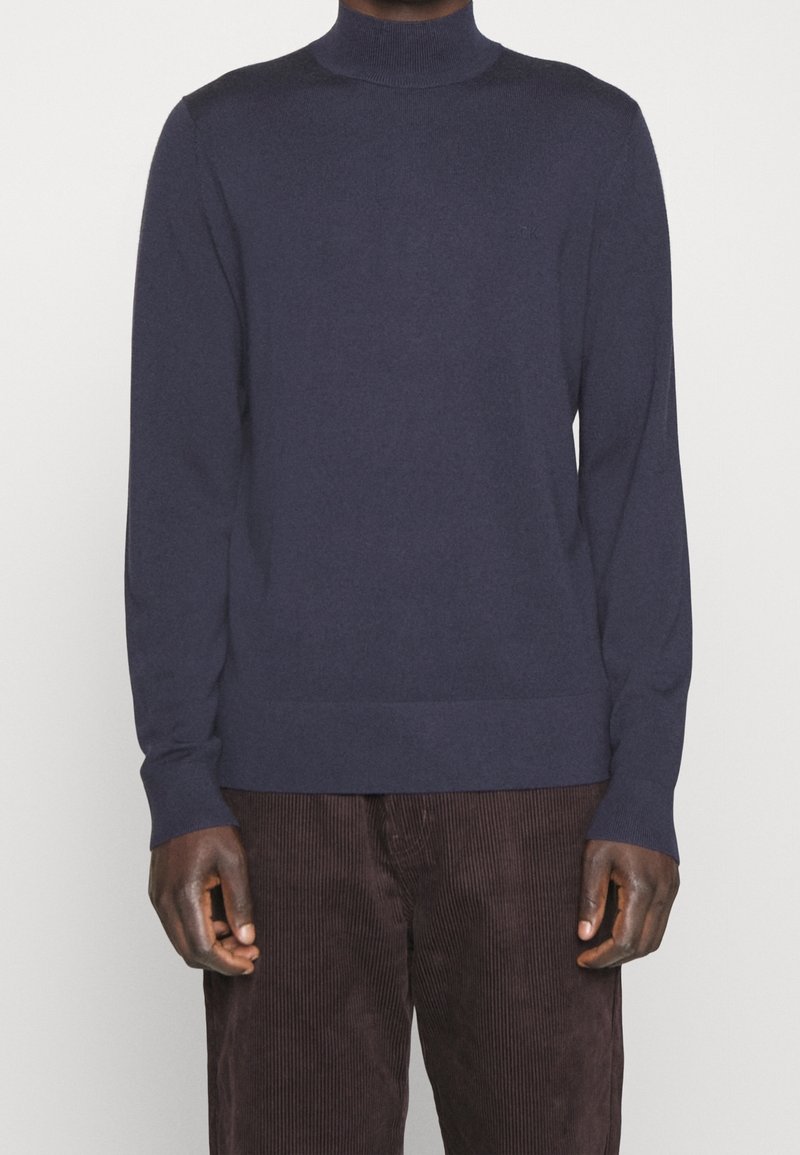 Homme portant un pull à col roulé bleu marine et un pantalon en velours côtelé marron foncé, debout devant un fond blanc.