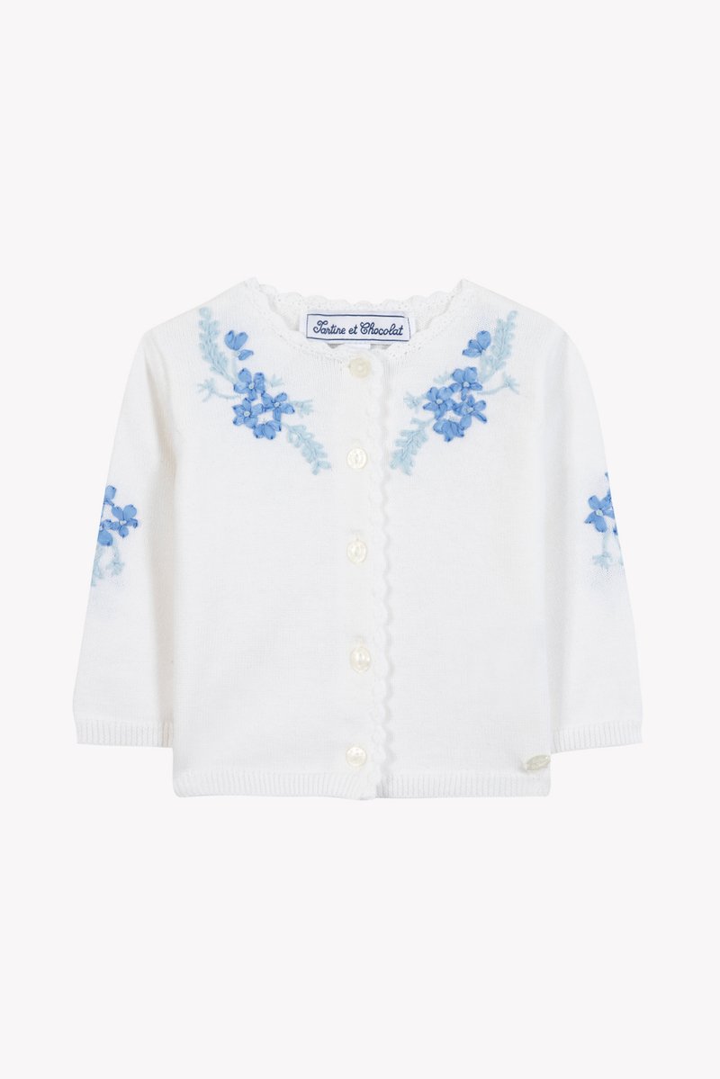 Λευκή πλεκτή ζακέτα με μακριά μανίκια, διακοσμημένη με μπλε φλοράλ κέντημα και στις δύο πλευρές και γιακά με κυματιστό τελείωμα. Περλέ κουμπιά κατά μήκος του μπροστινού μέρους.