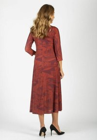 Kobieta w długim rękawie, w rdzawym, wzorzystym midi sukience i czarnych szpilkach, stoi plecami do aparatu.