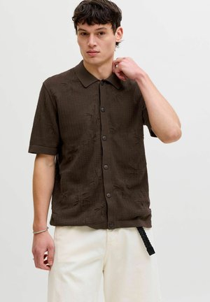 Młody mężczyzna poprawiający kołnierz, ubrany w teksturowaną ciemnobrązową koszulę na krótkim rękawie zapinaną na guziki oraz jasnobeżowe spodnie na jednolitym tle.