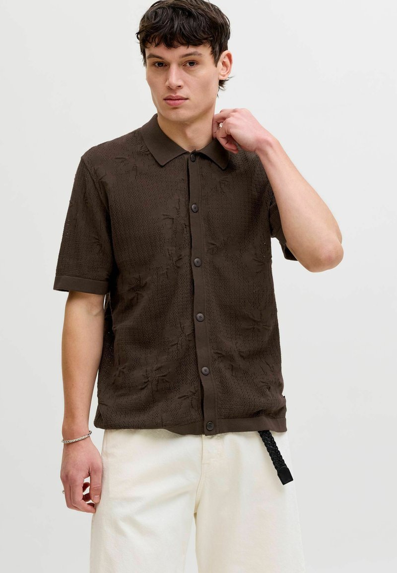 Jeune homme ajustant son col, portant une chemise marron foncé texturée à manches courtes avec boutons et un pantalon beige clair sur un fond uni.