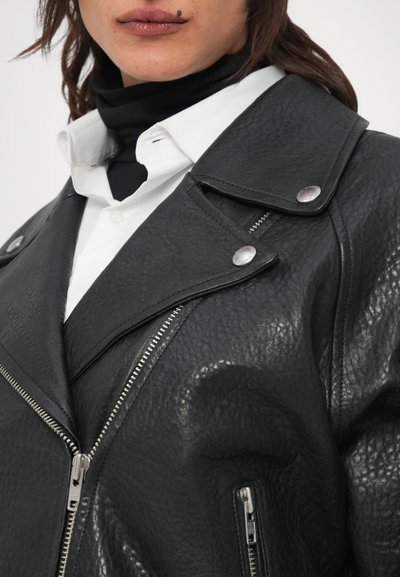 Casaco de motociclista em couro preto com acabamento texturizado, ferragens prateadas, lapelas largas e bolsos com fecho de correr, usado sobre uma camisa branca.