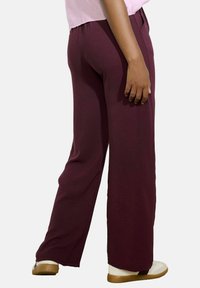 Burgundy kalhoty s širokými nohavicemi, vyrobené z měkké látky, s elastickým pasem, hladkou texturou a uvolněným střihem, doplněné béžovými teniskami.