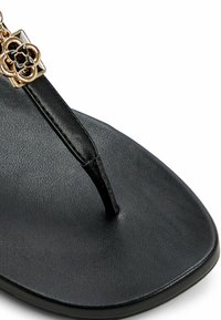 Black flip-flop sandal with a textured footbed and a T-strap featuring a gold floral accent. Smooth surface, minimalist design.