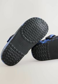 Chaussons bleus à motifs avec semelles en caoutchouc noir, dotés d'une texture antidérapante et de sangles ajustables sur le dessus.