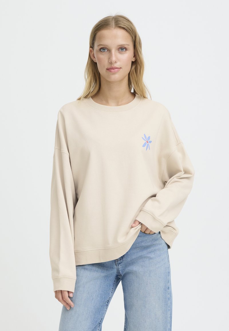 Sweat-shirt beige à coupe décontractée, encolure ronde et manches longues. Présente un petit motif floral bleu sur la poitrine.