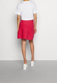 Person klädd i en vit t-shirt, ljusrosa kjol och vita plattformsskor som står mot en enkel ljus vägg och golv.