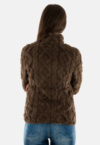 Giacca in pile marrone, texturizzata, con colletto alto e motivo a trecce. Abbinata a jeans in denim blu, vista da dietro.