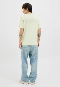 Lichtgele, t-shirt met korte mouwen en een ronde hals, gecombineerd met losse, lichtblauwe jeans. Opvallende tatoeages op de armen, natuurlijke stoftextuur.