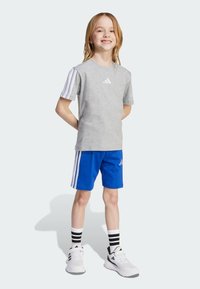 Camiseta gris de manga corta de Adidas con logo en blanco, combinada con pantalones cortos azules con rayas blancas. Calcetines de rayas en blanco y negro y zapatillas deportivas.