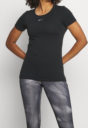 Femme portant un t-shirt noir ajusté Nike et des leggings gris à motifs, les mains posées sur les hanches devant un fond uni.