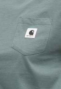 Türkis puuvillane T-särk, millel on rinnatasku valge sildiga, millel on must logo. Sile tekstuur ja kerge läige.