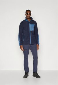 Polarowa kurtka w ciemnym granacie z jasnoniebieskimi akcentami, z kieszenią na piersi na zamek oraz pełnym zamkiem z przodu, w połączeniu z ciemnymi spodniami i okularami przeciwsłonecznymi.