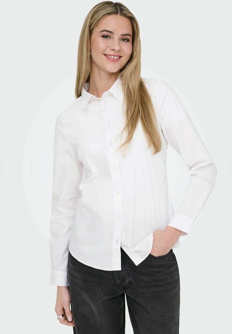 Jeune femme aux longs cheveux blonds portant une chemise blanche boutonnée et un jean noir, souriant avec une main dans la poche.
