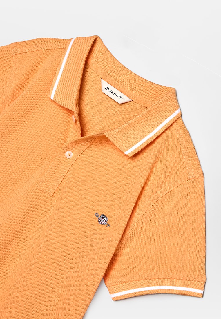 Polo orange avec des détails de col blanc, manches courtes, et un logo brodé sur la poitrine. La texture du tissu est côtelée.