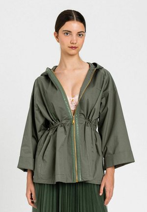 Giovane donna che indossa una giacca con cappuccio verde oliva aperta con vita aderente sopra un reggiseno in pizzo rosa chiaro e una gonna plissettata verde scuro.